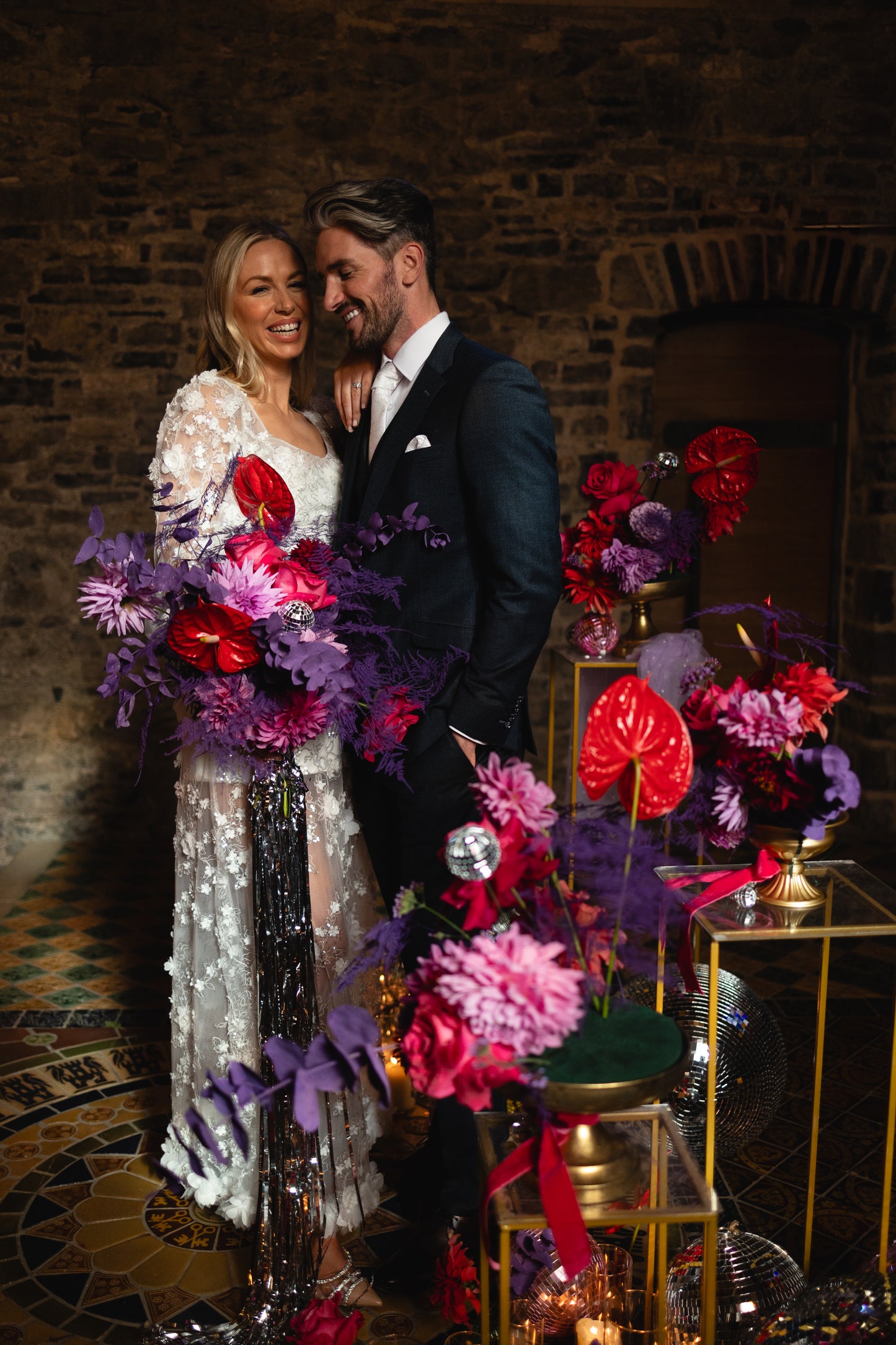 “Modern bride in a sheer floral appliqué wedding dress by Mizz Rio, posing against a stone wall in Dublin and holding a colourful bouquet – alternative bridal look for a city wedding