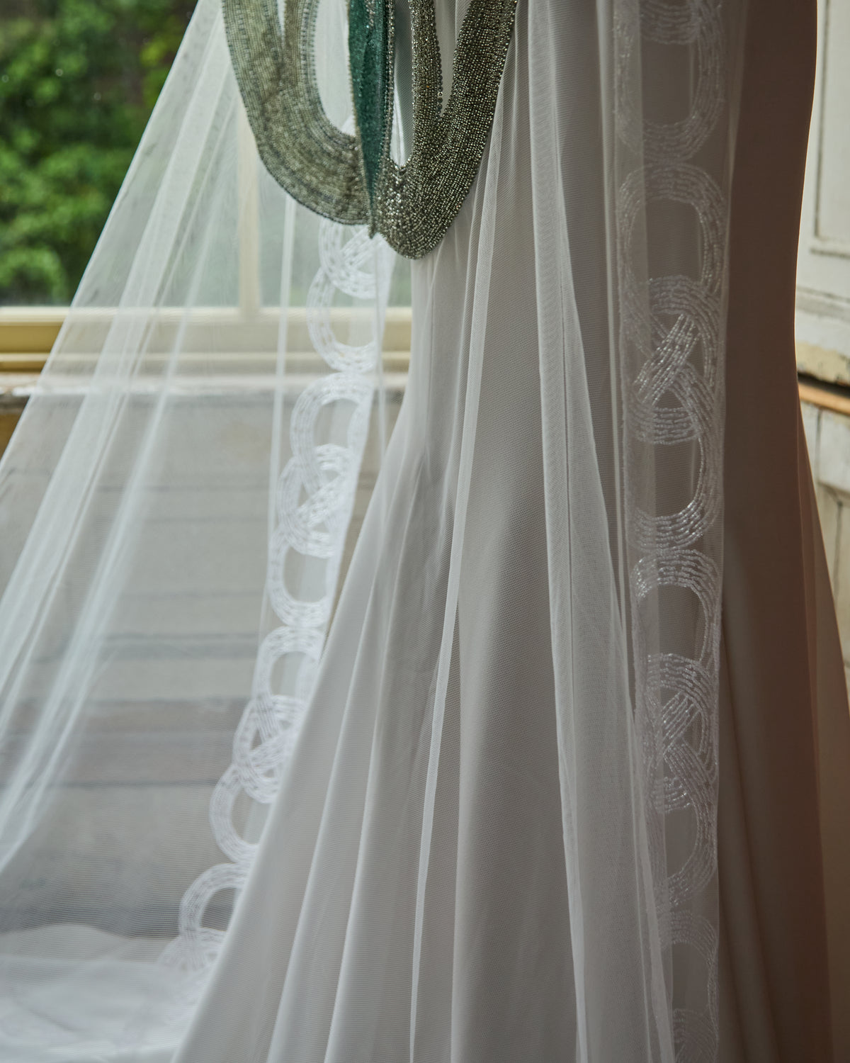 The Claddagh Couture veil, beaded with linked Claddagh hearts, hanging in window light