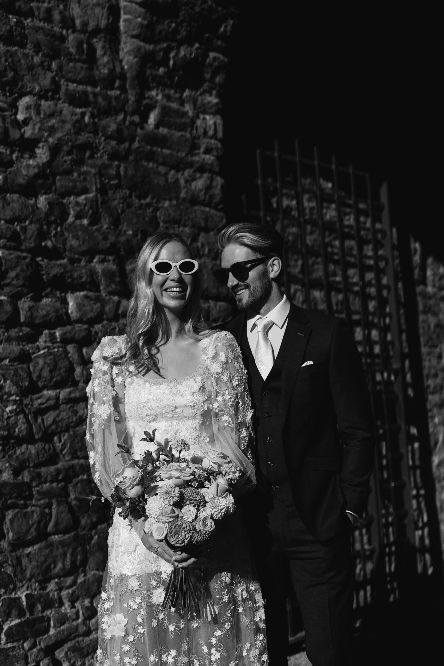 “Modern bride in a sheer floral appliqué wedding dress by Mizz Rio, posing against a stone wall in Dublin and holding a colourful bouquet – alternative bridal look for a city wedding