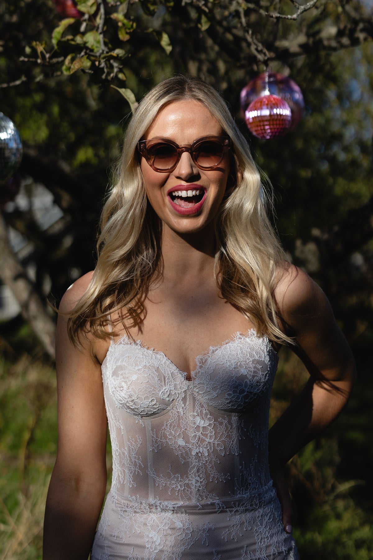 Laughing bride in the Fiadh Chantilly lace wedding dress with a sweetheart corset bodice, outdoors among disco balls – Mizz Rio